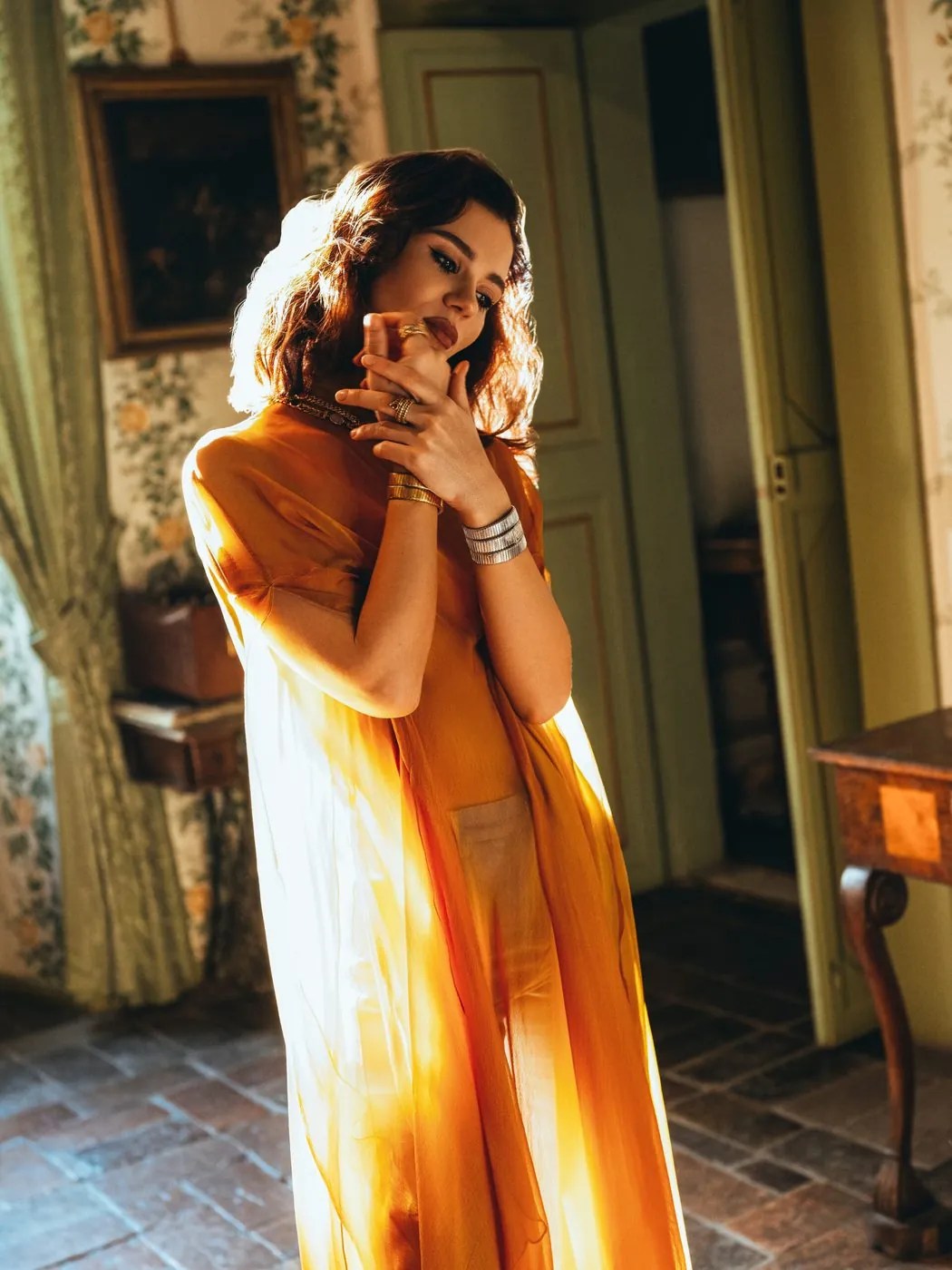 A model in an orange dress with bracelets and rings, backlit in a bedroom with rose wallpaper