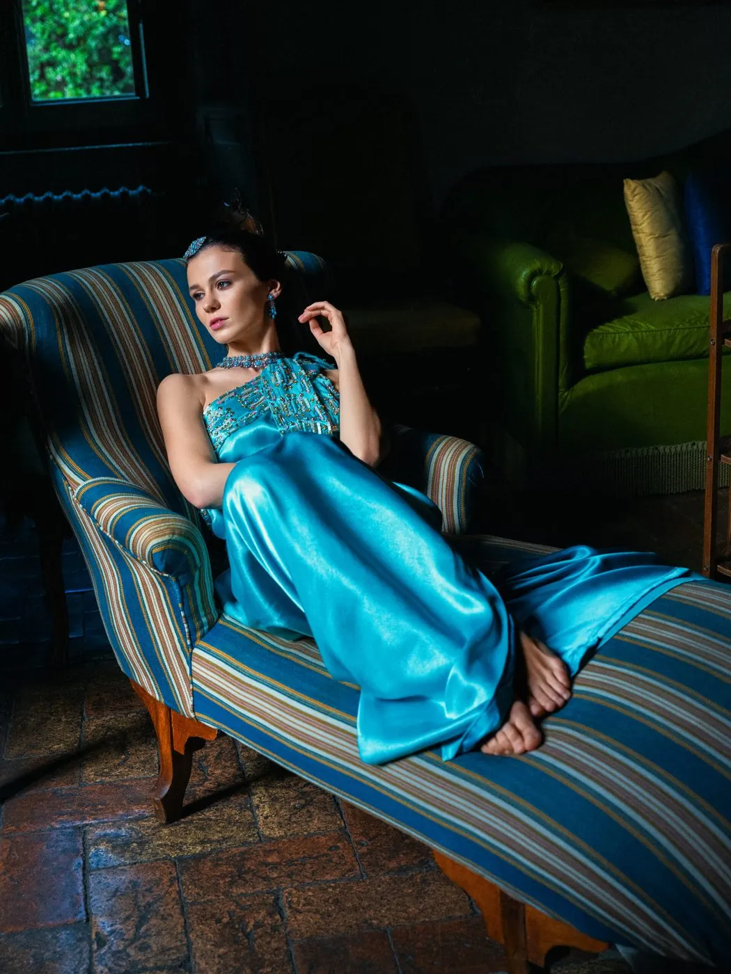 A model in a turquoise satin dress and jewellery, reclining on a striped chaise longue
