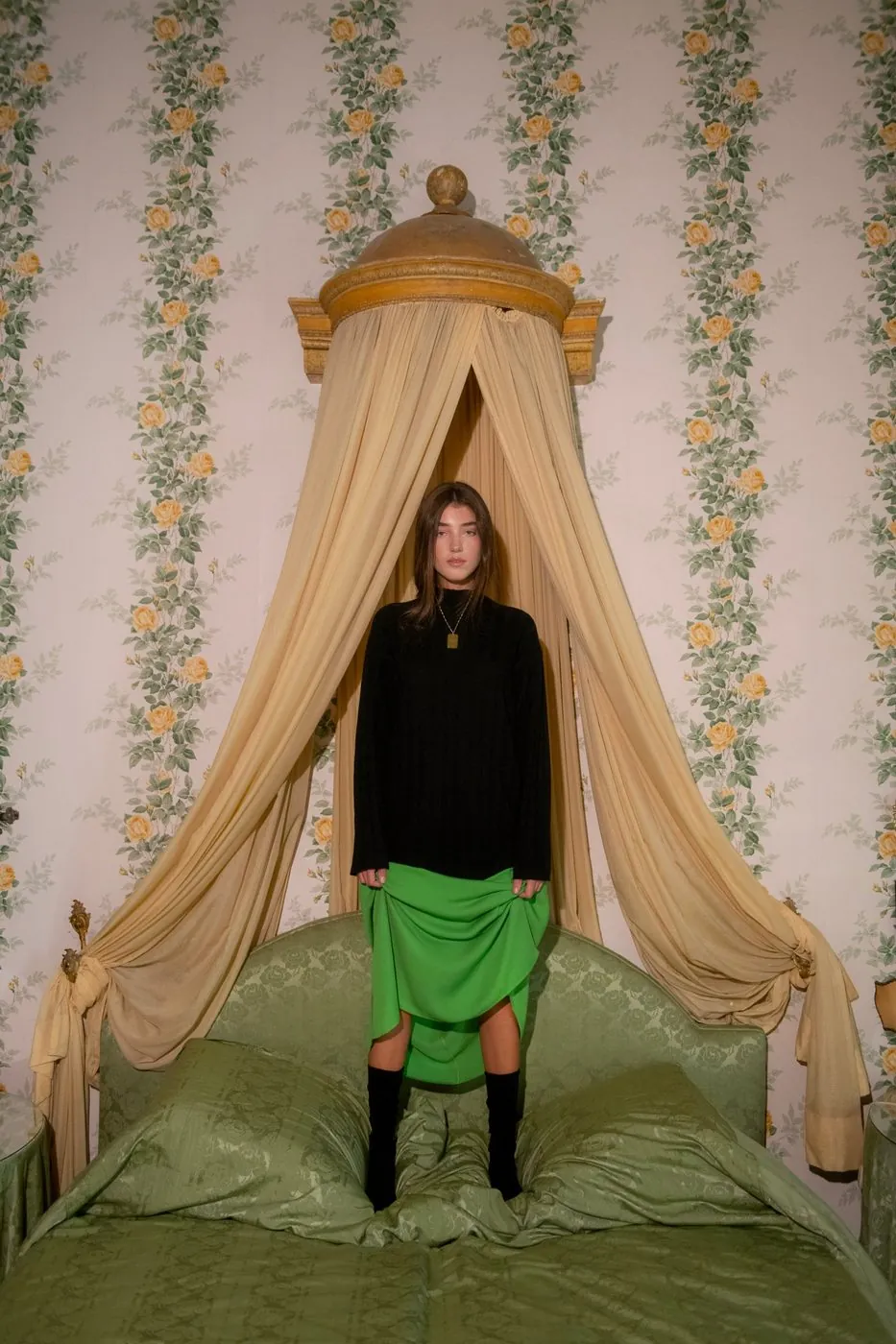 A model standing on a canopy bed, in a bedroom with rose wallpaper