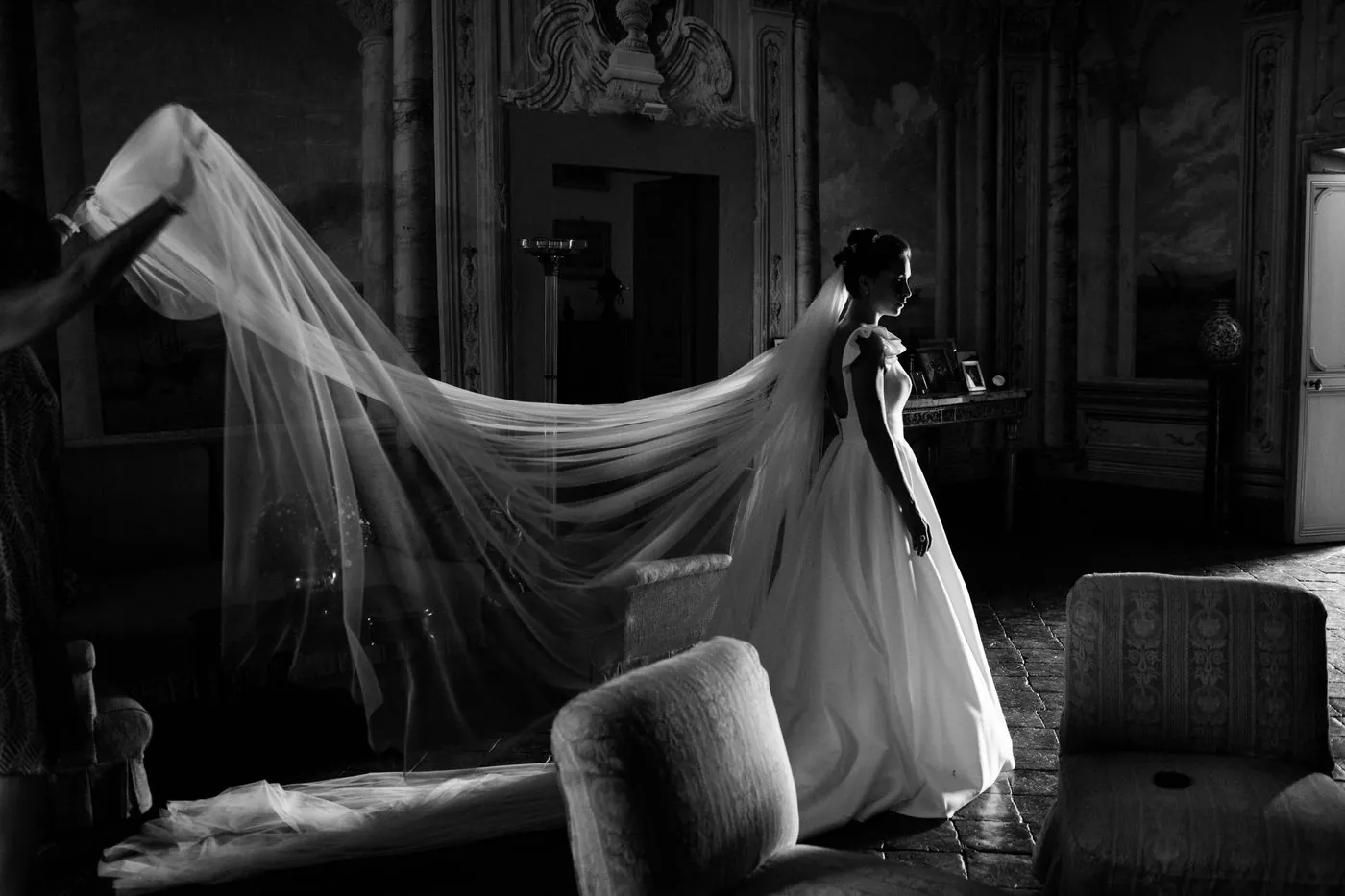 In black and white, the bride with her long veil lifted in the frescoed Gallery